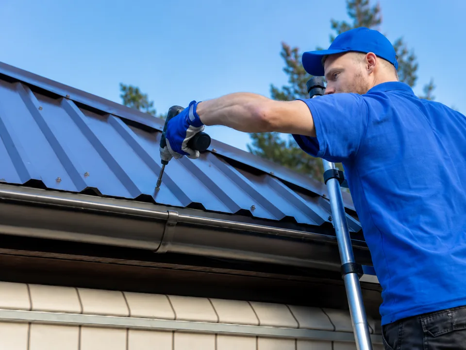 Roofer on a ladder applying sealant along the eave of a dark blue standing seam metal roof
