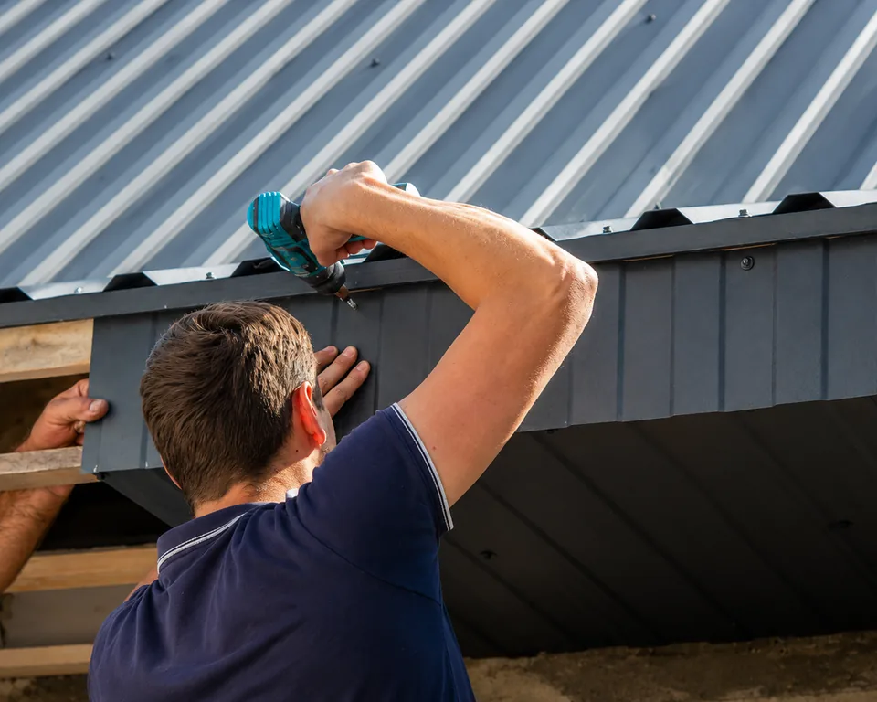 Close-up of a worker refitting the edge trim beneath the eave of a grey metal roof
